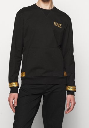 Schwarzer Sweatshirt mit langen Ärmeln und goldenen Emporio Armani EA7 Logos auf der Brust, an den Bündchen und in der Nähe des Saums, getragen von einer stehenden Person.