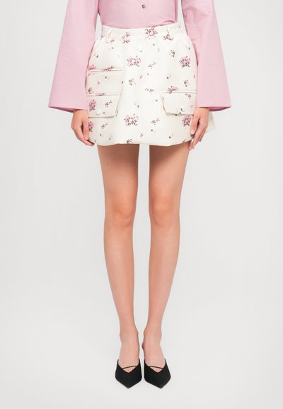 Jupe mini en tissu crème à motifs floraux, avec deux poches avant, une taille élastique et une coupe droite, associée à des escarpins noirs à bout pointu.
