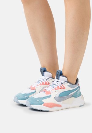 Nogi w białych, niebieskich i koralowych sneakersach Puma RS, stojące na jednolitym białym tle.