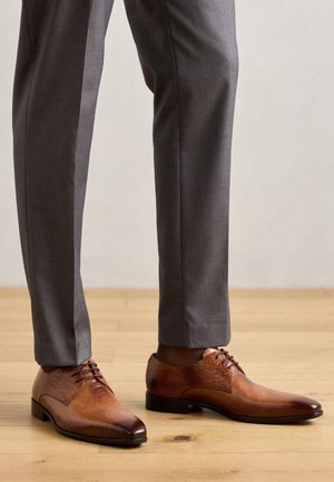 Pantofi de piele maro cu un finisaj texturat, vârful ascuțit și șireturi, purtați împreună cu pantaloni gri suitați pe un podea din lemn.