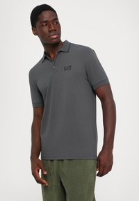 Man wearing dark gray polo shirt with EA7 logo and textured green pants, looking to his left against a plain white background.