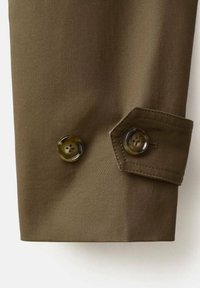 Gros plan sur un poignet de manche en tissu marron avec deux boutons marron à quatre trous, l'un sur un détail de bride et l'autre fixé directement à la manche.