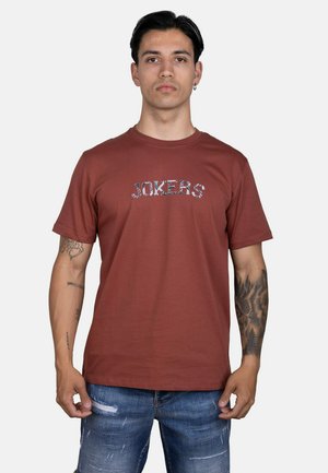 Hombre con cabello corto y oscuro y tatuajes lleva una camiseta color óxido con el texto "JOKERS" y vaqueros azules rotos, de pie frente a un fondo sencillo.