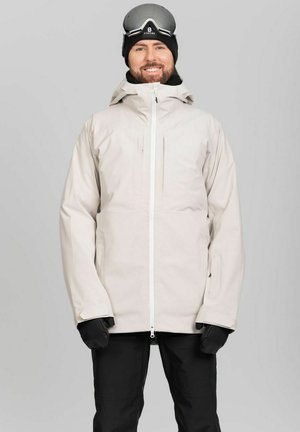 Mand iført hvid vinterjakke, sorte handsker, sorte bukser og sort hue med skibriller på toppen, smilende mod grå baggrund.