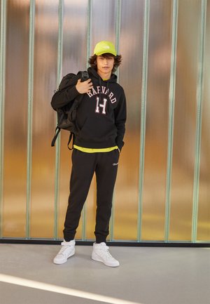 Felpa nera con testo "HARVARD", maglietta gialla sotto, pantaloni sportivi neri, scarpe bianche e un cappellino verde neon. Zaino sulla spalla sinistra.