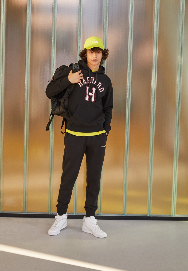 Felpa nera con testo "HARVARD", maglietta gialla sotto, pantaloni sportivi neri, scarpe bianche e un cappellino verde neon. Zaino sulla spalla sinistra.