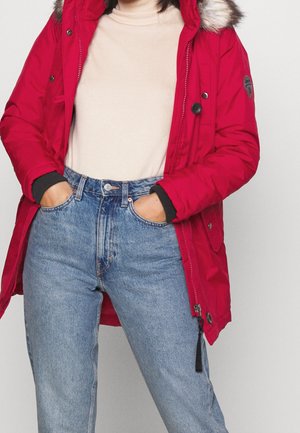 Femme portant un parka rouge avec une capuche bordée de fourrure, un pull à col roulé beige et un jean bleu taille haute, les mains dans les poches.
