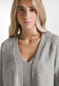 Grau gestrickte Strickjacke mit tiefem V-Ausschnitt und gerippter Struktur über einem passenden grauen Oberteil. Weicher Stoff mit leicht lockerer Passform.
