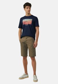 Marineblaue T-Shirt mit einer Landschaftsgrafik, kombiniert mit khakifarbener Shorts. Die Sneakers sind weiß mit beigen Akzenten. Lässiges Sommeroutfit.