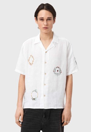 Ung person med kort mørkt hår som bruker en hvit kortermet skjorte med blomsterbroderi og mørke bukser.