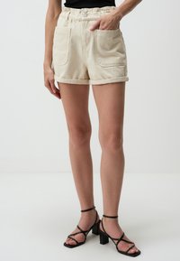 Shorts di denim beige con vita elastica, due tasche frontali e orli arrotolati. Abbinati a sandali con tacco a strappi neri.