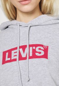 Felpa grigia con cappuccio e logo a scatola rosso con la scritta "LEVI'S" in bianco. Presenta lacci e un tessuto morbido e texturizzato. Senza motivi o accenti.