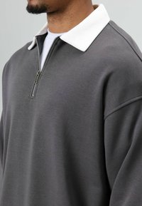 Grijze trui met een witte kraag, voorzien van een ritssluiting bij de hals, zachte textuur en een ontwerp met verlaagde schouders. Close-up weergave.