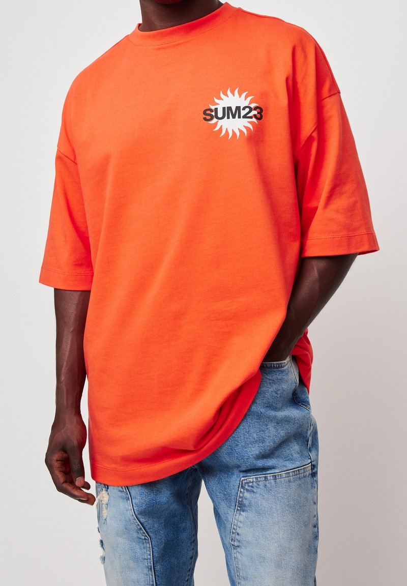 T-shirt en coton orange vif avec un col rond, coupe oversize, et un graphisme noir "SUM23" entouré d'un motif rayonnant sur le devant.