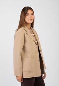 Blazer in misto lana beige con colletto a rever, bottoni a un solo petto e due tasche frontali. Tessuto morbido e vestibilità sartoriale.