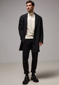 Manteau en laine gris foncé avec une coupe décontractée, associé à un pull à col roulé crème et un pantalon noir, complété par des chaussures noires à lacets.