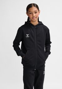 Schwarze Zip-Hoodie aus weichem Material, mit Kapuze, Kängurutaschen und Logo auf der linken Brust. Kombiniert mit passenden Jogginghosen.