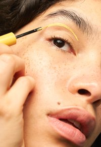 Gelbe Eyeliner wird entlang der Braue aufgetragen. Die Person hat einen glatten Teint mit Sommersprossen, dunklem Haar und natürlicher Lippenfarbe.