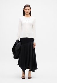 Ein weißes Langarmshirt, kombiniert mit einem schwarzen asymmetrischen Rock. Das Outfit wird durch eine silberne Halskette und schwarze Absatzsandalen ergänzt.