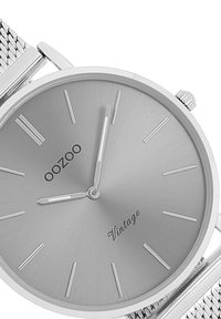 Silver stainless steel watch with a minimalist design, featuring a smooth gray dial, slender hour markers, and mesh strap. Brand logo visible.