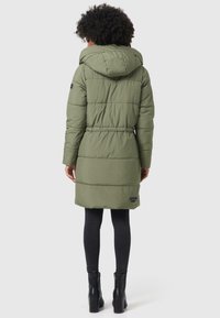 Olijfgroene gevoerde jas met capuchon, voorzien van een elastische taille, lange mouwen en een logo patch vlakbij de zoom, gecombineerd met zwarte skinny jeans en laarzen.