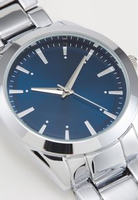 Montre en acier inoxydable argenté avec un cadran texturé bleu, des index en argent et des aiguilles lumineuses. Équipée d'un bracelet en métal poli.