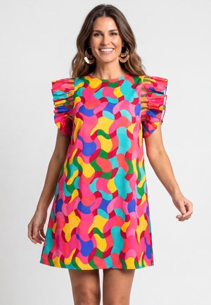 Frau lächelnd, trägt ein buntes kurzes Kleid mit abstrakten wellenförmigen Mustern und kurzen gerafften Ärmeln, steht vor einem einfarbigen hellen Hintergrund.