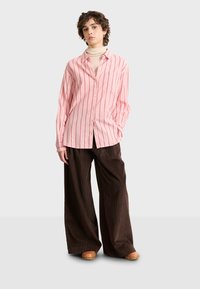 Light pink button-up shirt with vertical red stripes, paired with wide brown corduroy pants. Smooth texture and relaxed fit.