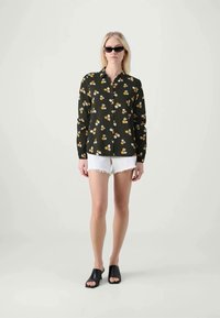 Svart blommig blus med vita och gula blommor, ihopparad med vita denimshorts. Modell som bär svarta slip-on sandaler och solglasögon.