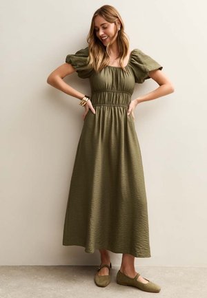 Vrouw lacht met handen in de zij, draagt een olijfgroene midi-jurk met pofmouwen en bijpassende Mary Jane ballerina's, tegen een effen achtergrond.