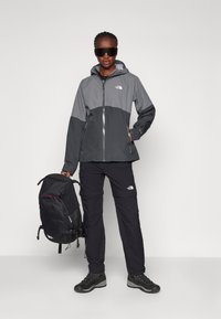 Geacă waterproof gri și neagră cu glugă, asortată cu pantaloni negri și bocanci de drumeție. Modelul poartă un rucsac negru.