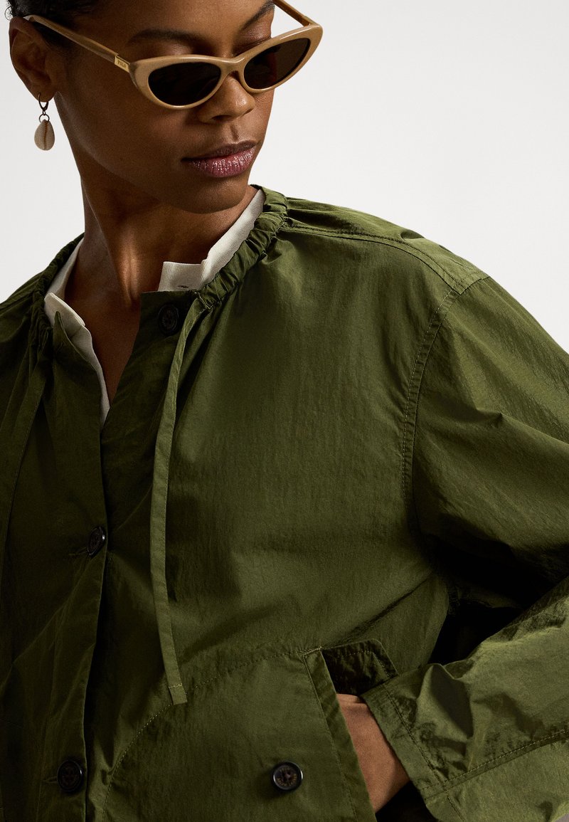 Woman wearing olive green buttoned jacket, beige cat-eye sunglasses, and shell drop earrings, posing with hand in jacket pocket.