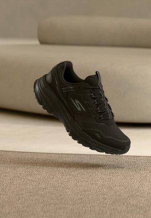 Μαύρο αθλητικό παπούτσι με υφή στην σόλα και κορδόνια, με ανακλαστικό λογότυπο "S", τοποθετημένο πάνω σε μπεζ χαλί και φόντο με καμπύλο καναπέ.