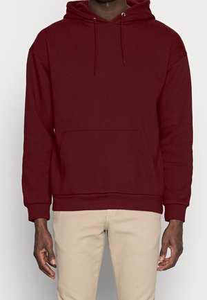 Mann trägt einen schlichten burgunderroten Hoodie mit Fronttasche und beige Hose, steht vor einem hellen Hintergrund.