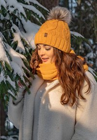 Žlut�ý pletený čepice s kožešinovým pom-pom a odpovídajícím pleteným šál. Měkká, texturovaná tkanina. Modelka nosí bílý fleecový kabát v zasněženém prostředí.
