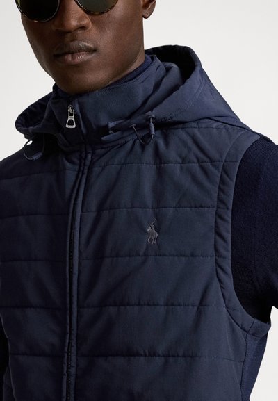 Vyras dėvi tamsiai mėlyną dygsniuotą liemenę su gaubtu ir juodus saulės akinius, apkarpyta nuo pečių iki smakro lygio.