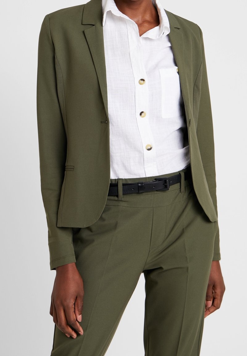 Blazer vert olive et pantalon assorti, coupe ajustée, fabriqués en tissu lisse. Chemise blanche avec boutons et poche poitrine. Ceinture noire en accent.