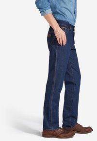 Jeans de algodón azul oscuro con corte recto, con costuras en color naranja y un parche de cuero marrón; combinados con una camisa de botones azul claro.