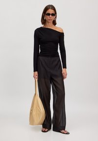 Zwarte top met één schouder, gecombineerd met zwarte doorzichtige wijde broek. Het model houdt een beige leren tote bag vast en draagt zwarte zonnebrillen en sandalen.
