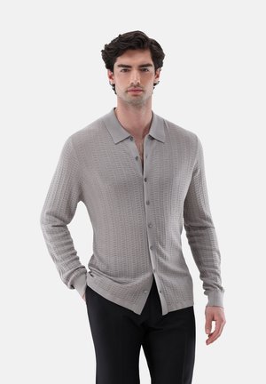 Mann trägt eine hellgraue, strukturierte Knopf-Cardigan mit Kragen und schwarze Hose, steht mit einer Hand in der Tasche vor einfarbigem Hintergrund.