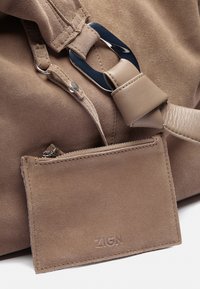 Beżowa torebka z zamszu z gładkim skórzanym paskiem, metalowymi akcentami oraz dołączonym etui na karty z zamkiem błyskawicznym i wytłoczonym logo.