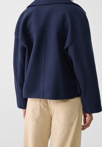 Marinblå ullblandad jacka med vida ärmar, strukturerade axlar och kort kirt, i kombination med beige högmidjade jeans.