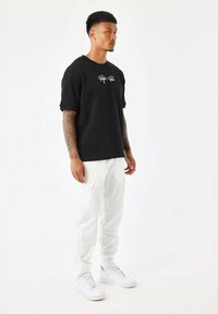 Camiseta negra de gran tamaño con logo, pantalones cargo blancos y zapatillas blancas. El conjunto cuenta con mangas cortas y bolsillos cargo en los pantalones.
