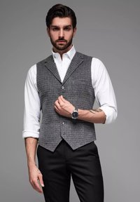 Grijze geruite vest met een V-hals, gecombineerd met een wit shirt. Inclusief knopen en een borstzak. Man draagt een horloge om zijn linker pols.