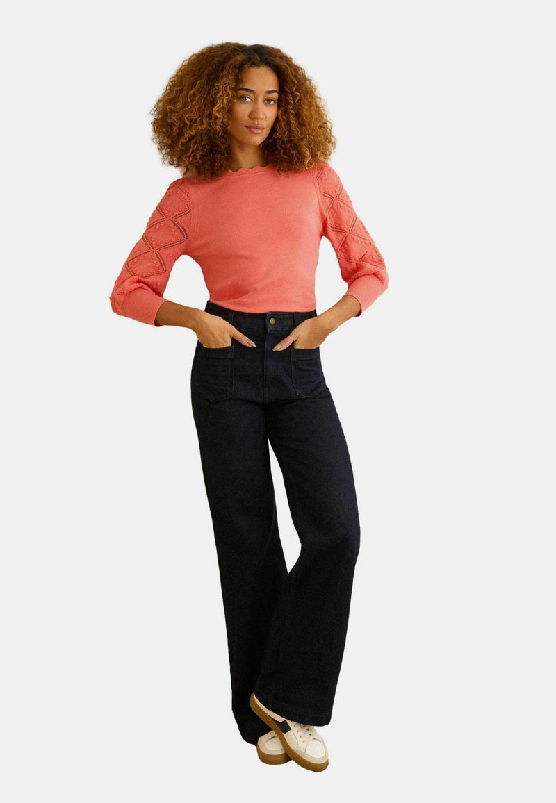 Vrouw met krullend haar, gekleed in een koraalkleurige gebreide top met gestructureerde mouwen, een donkere, hooggetailleerde spijkerbroek en witte sneakers, staand met handen in de zakken.