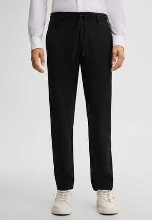 Mann trägt schwarze Kordelhose mit weißen Sneakers und weißem Langarmshirt, steht vor einem schlichten Hintergrund.