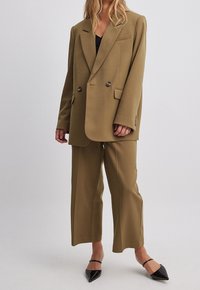 Podwójnie zapięta oliwkowo-zielona marynarka z wyrazistymi klapami, w połączeniu z dopasowanymi spodniami o szerokich nogawkach; materiał wydaje się gładki i dopasowany, dopełniony spiczastymi czarnymi szpilkami.