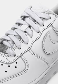 White leather sneaker with grey accents, padded tongue, round laces, and perforations. Features a textured Nike logo and contrasting stitching.