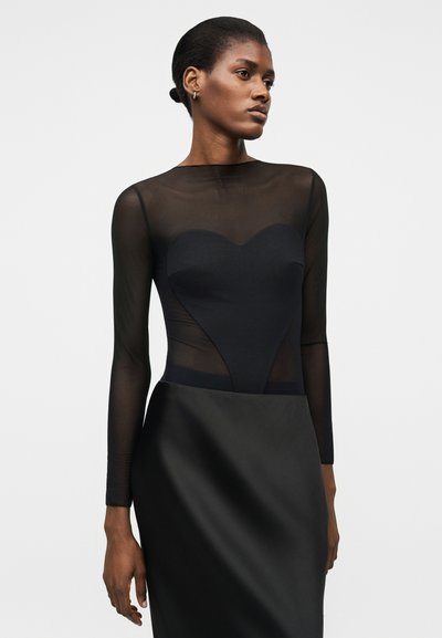 Γυναίκα με μαύρο διαφανές τοπ μακρυμάνικο με καρδιοειδές πάνελ και μαύρη σατέν φούστα, κοιτάζοντας στο πλάι με φόντο ένα λευκό υπόβαθρο.