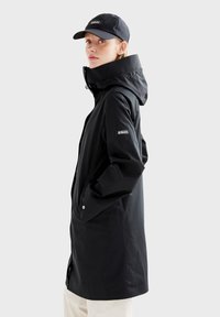 Veste imperméable noire avec un col montant, des poches latérales et un logo sur la manche. Associée à une casquette noire assortie.
